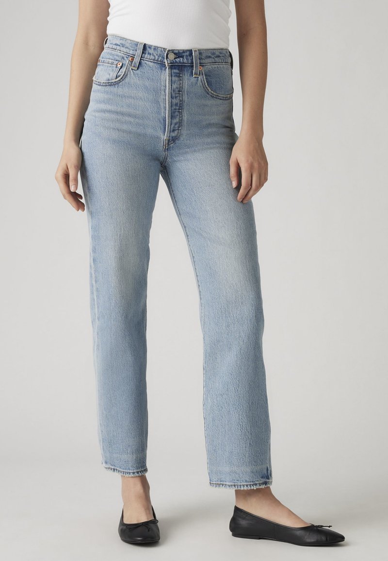 Femme portant un jean bleu clair taille haute avec des jambes droites et des ballerines noires, debout devant un fond clair uni.