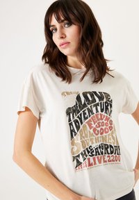 Camiseta de algodón crema con mangas cortas, que presenta una tipografía en negrita y texturizada en negro, dorado y rojo, centrada en el frente.