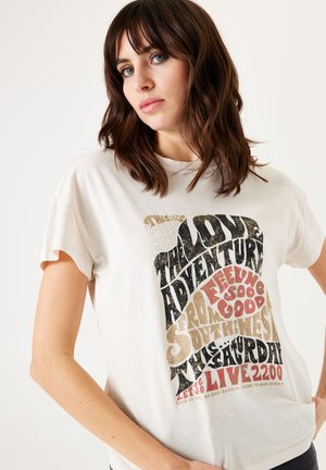 T-shirt en coton crème à manches courtes, présentant une typographie audacieuse et texturée en noir, or et rouge, centrée sur le devant.