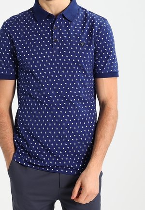 Man die een marineblauw poloshirt met een klein wit palmboompatroon en donkerblauwe broek draagt, staand met één hand in zijn zak.