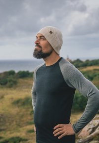 Gebreide roomkleurige muts met gestructureerd patroon; donker shirt met lange mouwen en grijze mouwen; buitenomgeving met groen en oceaan op de achtergrond.