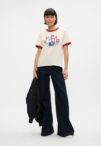 Κρέμα κοντομάνικη μπλουζάκι με μπορντώ γιακά, με εκτύπωση γραφικών που περιλαμβάνουν το κείμενο "CUPID LUCKY YOU." Φορεμένο με σκούρες φαρδιές τζιν.