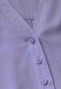 Blusa viola chiaro con scollatura a V, tessuto morbido e cinque bottoni coordinati lungo la pattina anteriore.