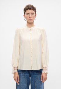 Donna con capelli corti che indossa una blusa lunga a maniche con pizzo ai polsini e dettagli dei bottoni, abbinata a jeans blu, in piedi contro uno sfondo bianco.