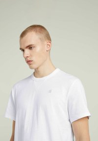 T-shirt bianco a maniche corte con colletto girocollo e un branding discreto sul petto, realizzato in tessuto leggero e morbido con una texture liscia.
