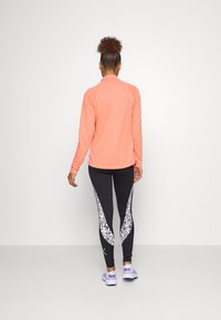 Veste de sport légère à manches longues couleur pêche, associée à des leggings noirs avec des panneaux latéraux floraux blancs et des chaussures de sport.