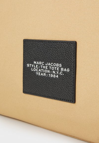 Musta tekstuuriga silt beežist kangast käekotis, millel valge tekst, mis kannab nime Marc Jacobs, käekoti stiil, New Yorgi asukoht ja aasta 1984.