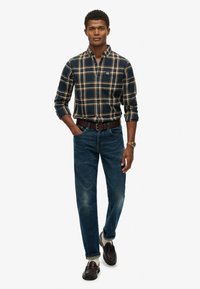 Camisa de cuadros en navy y marrón claro, combinada con jeans azules. Cinturón marrón y mocasines oscuros completan el conjunto. Estilo casual y entallado.