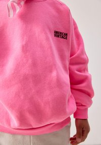 Person trägt ein leuchtend pinkes Sweatshirt mit dem Schriftzug "American Vintage" auf der Brust und beigefarbene Hosen, Arm entspannt an der Seite.