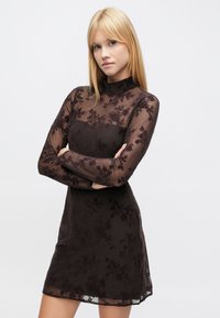 Robe marron transparente à manches longues, col haut, motif en dentelle florale, et corsage ajusté, se terminant au-dessus du genou. Texture lisse.