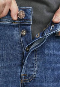 Jeans in denim blu con una vista ravvicinata della vita, caratterizzati da un bottone in rame, accessori in ottone e dettagli di cucitura a contrasto.