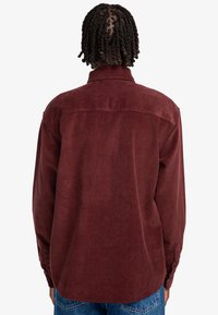 Camisa de pana burdeos con mangas largas, puños de botón y detalle en el yugo trasero. Textura suave y ajuste relajado.