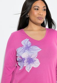 Kvinde iført magentarød langærmet skjorte med lilla blomsterdesign på brystet, kigger let mod højre, med creoløreringe og glat mørkt hår.