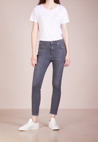 Jeans ajustados de talle alto grises con diseño de cinco bolsillos, combinados con una camiseta blanca de cuello redondo y zapatillas blancas, sobre un fondo neutral.