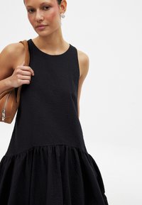 Musta ilmanhiha dressi, jossa on teksturoitu yläosa ja flarettava, poimutettu helma. Malli pitää kädessään vaaleanruskeaa käsilaukkua, mikä tuo esille hienovaraisia kangasdetaljeja.