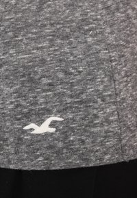 Gråmelerad bomullst-shirt med en kort svart kant och en vit broderad fågel-logotyp placerad nära nederkanten till vänster.