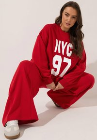 Sudadera roja de gran tamaño con "NYC 97" en texto blanco, combinada con pantalones de pierna ancha a juego. Zapatillas blancas con acentos grises.