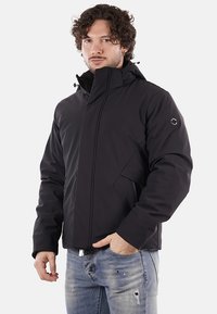 Uomo con capelli ricci che indossa una giacca a vento nera con cappuccio e jeans blu strappati, in piedi con una mano in tasca su uno sfondo semplice.