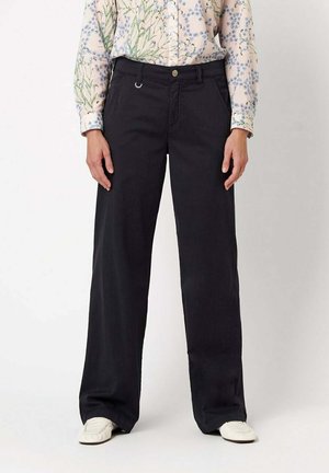 Person trägt hoch taillierte schwarze weite Hose, geblümte Langarmbluse, die in die Hose gesteckt ist, und weiße flache Schuhe, steht vor weißem Hintergrund.