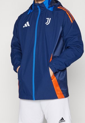Mężczyzna ubrany w niebiesko-pomarańczową sportową kurtkę Juventusu z białymi szortami Adidas, ręce w kieszeniach kurtki, stoi na jednolitym tle.