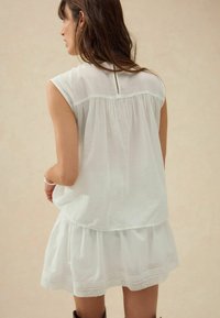 Robe blanche avec un design en couches, présentant un dos froncé, des manches courtes, et une bordure en dentelle le long de l'ourlet, fabriquée en tissu léger.