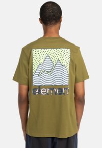 Olivegrünes Baumwoll-T-Shirt mit einem großen Grafikdesign von stilisierten Bergen mit wellenförmigen Linien und Punkten auf einem hellgrünen Hintergrund. Logo darunter.