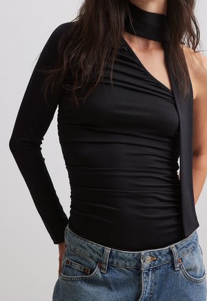 Femme portant un haut noir à une manche, avec un détail de nœud au cou et un jean bleu, debout les mains dans les poches ; tête partiellement hors du cadre.