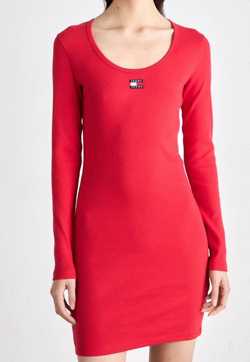Femme portant une robe rouge ajustée à manches longues avec un décolleté en rond et un petit logo Tommy Jeans sur la poitrine.