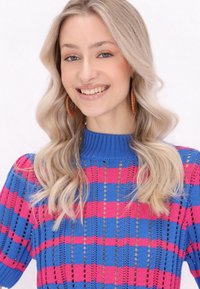 Sweter z dzianiny w niebiesko-różowe kratki, z prążkowanym dekoltem i otwartą koronkową fakturą. Modelka uśmiecha się, pokazując kolczyki w kształcie kół.