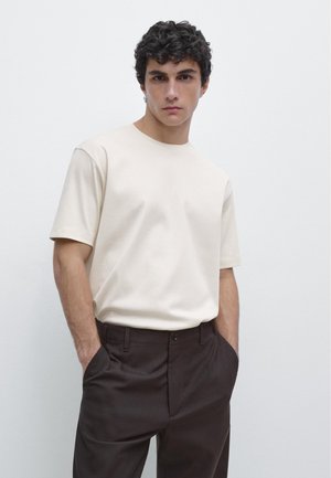 Giovane uomo con capelli ricci e scuri che indossa una maglietta beige a maniche corte senza disegni e pantaloni marrone scuro, con le mani in tasca, sfondo neutro.