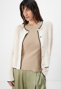 Cardigan bianco a maglia con texture e bottoni neri, indossato sopra una maglietta beige a costine. Abbinato a pantaloni larghi di un verde oliva chiaro.
