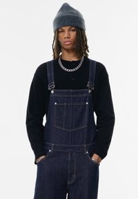 Salopette in denim con bretelle regolabili, tasca anteriore e lavaggio scuro. Abbinata a una maglietta nera a maniche lunghe e un berretto in maglia grigio.