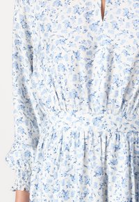 Vestido floral en azul claro y blanco, con escote en V, cintura fruncida, mangas largas con puños fruncidos y una tela suave y ligera.