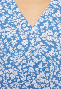 Robe florale bleue avec un décolleté en V, ornée de motifs de fleurs blanches et de cinq boutons blancs sur le devant. Tissu texturé.