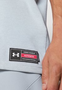 Camiseta deportiva de color azul claro con una tela texturizada, que presenta una etiqueta de "Under Armour" en negro y rojo que indica "AUTÉNTICO" y garantía de rendimiento.