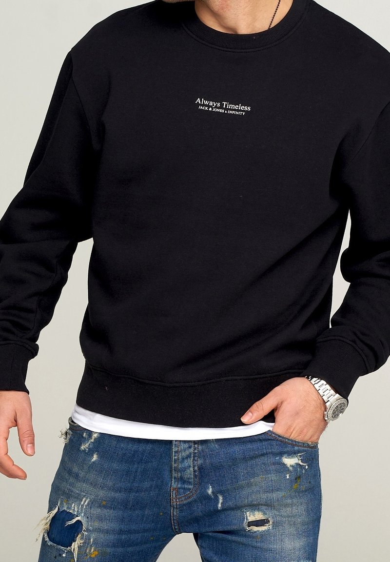 Homme portant un sweat-shirt noir avec le texte « Always Timeless Jack & Jones x Infinity » et un jean bleu déchiré, main dans la poche, montre argentée visible.