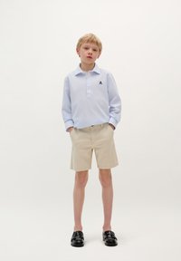 Camicia azzurro chiaro a maniche lunghe con un motivo testurizzato e patta dei bottoni, abbinata a pantaloni corti beige e mocassini neri.