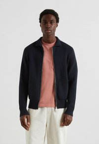 Giacca a maglia blu navy con zip, colletto a coste, due tasche frontali e tessuto a trama, indossata sopra una camicia rosa e pantaloni chiari.