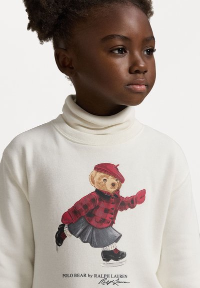 Balts turtlenecks džemperis ar grafiku, kas attēlo lāci sarkānā beretē, ar raibu žaketi un svārkiem, uz ledus slidojot ar melniem akcentiem.