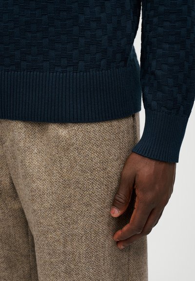 Dunkelblauer, strukturierter Pullover mit geripptem Saum und Bündchen. Hellbraune Tweed-Hose mit einem subtilen Muster. Eine sichtbare Hand liegt auf der Hose.