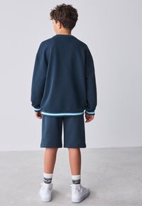 Marineblå sweater med tekstureret stof og blå stribede accenter, kombineret med matchende shorts. Hvide sneakers og ankelhøje sokker fuldender looket.
