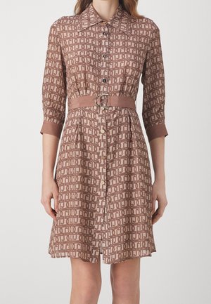 Frau trägt ein knielanges Kleid mit beigem und braunem Muster, einem Gürtel in der Taille und Dreiviertelärmeln.