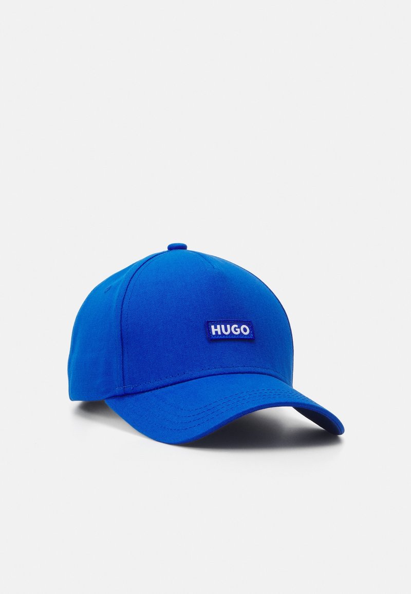 Casquette de baseball bleu vif avec une visière incurvée et un patch logo "HUGO" au centre devant, sur un fond blanc uni.