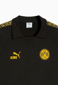 Sort polo-shirt med gule detaljer. Har krave, "KING" trykt i gult og et rundt logo med "BVB 09." Dekorativt mønster på skuldrene.
