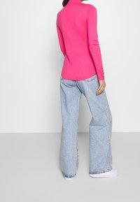 Suéter rosa ajustado con cuello alto y mangas largas, con puños abotonados, combinado con jeans de pierna ancha de color azul claro y zapatillas blancas.