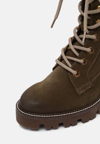 Brauner Wildlederstiefel mit runder Zehenpartie, kontrastierenden weißen Nähten, bronzenen Beschlägen und geflochtenen Schnürsenkeln. Verfügt über eine robuste, strukturierte Sohle.