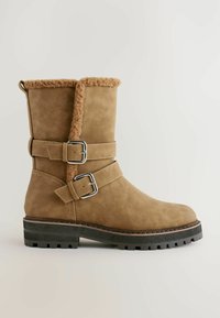 Tan ruskinds ankelstøvle med en fluffy kant, to sølvspændte remme og en robust sort gummisål med et tekstureret slidbanemønster.