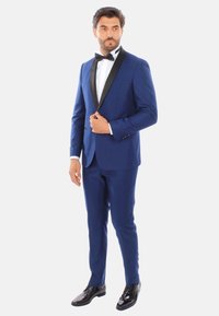 Tuxedo blu royal con bavero sciallato nero, camicia bianca e papillon. Design aderente, tessuto liscio, silhouette su misura, scarpe nere lucide.