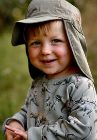 Dítě oblečené v světlé šedé mikině s potiskem dinosaurů, doplněné zeleným slunečním kloboukem. V pozadí je rozmazaná zeleň.