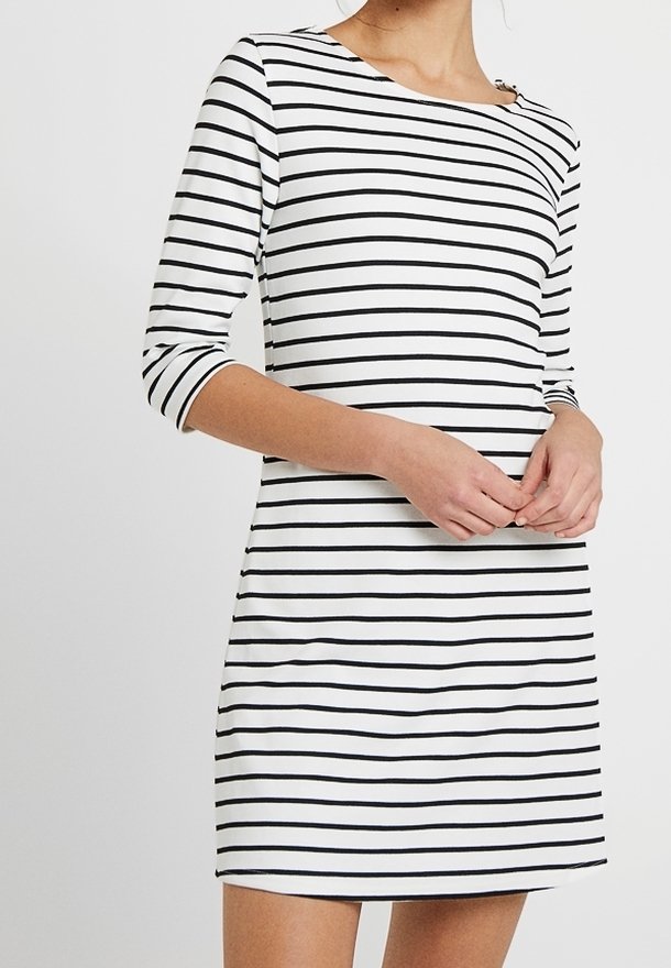 Femme portant une robe blanche à rayures horizontales noires et manches trois-quarts, mains jointes devant, sur fond uni.